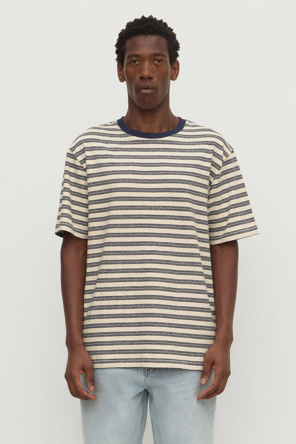 A man wearing a navy and cream short sleeve stripe tee with light blue washed denim jeans, standing against a plain white background.