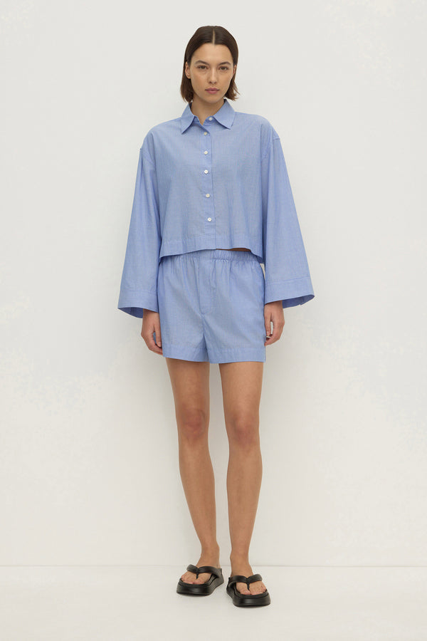A woman wearing a blue and white boxer style mini short, paired with a matching cropped shirt, and padded leather thongs, against a white background.